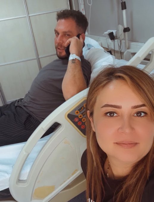 A man rests in a hospital bed talking on his phone while a woman takes a selfie in the foreground, with the medical bed's control panel showing orange buttons visible between them. The image captures the couple's bond during the man's hospitalization, aligning with the post's gratitude toward his supportive spouse just before his expected discharge.