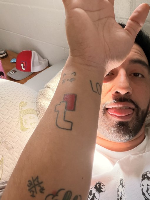 A man holds up his arm to show a tattoo of the TikTok logo alongside other tattoos, including one that appears to be a sad cat face.