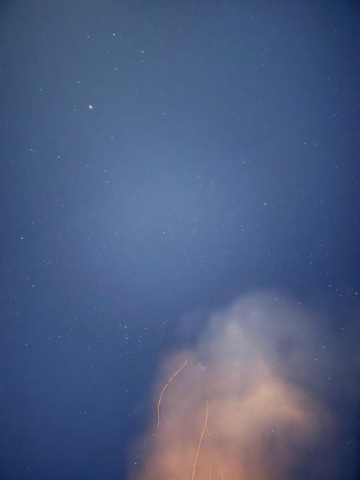 A twilight photograph of a starry blue sky with a bright celestial body visible, alongside a plume of smoke and glowing sparks rising from the bottom of the frame.
