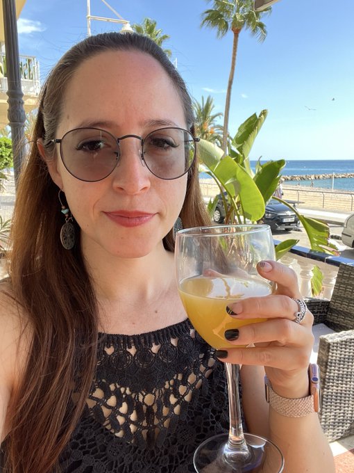 A selfie of a woman with glasses and a black crocheted top holding a glass of orange juice, taken at an outdoor seaside restaurant with palm trees and a parked car in the background.