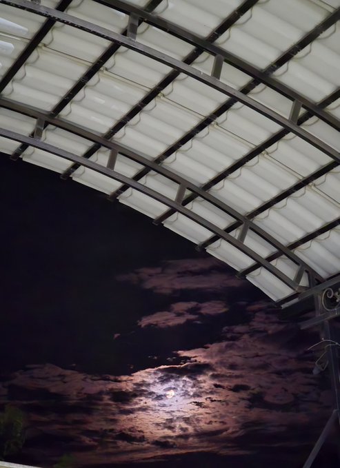 A nighttime photograph looking up at a corrugated metal roof structure with the bright moon partially visible through clouds in the dark sky below it.