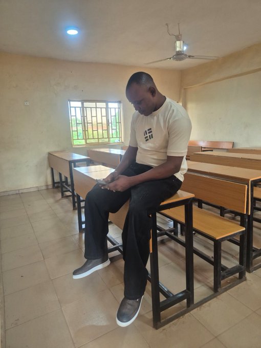 A photograph of a young man wearing a cream-colored shirt and dark pants sitting on a desk in a classroom, looking down at his phone.