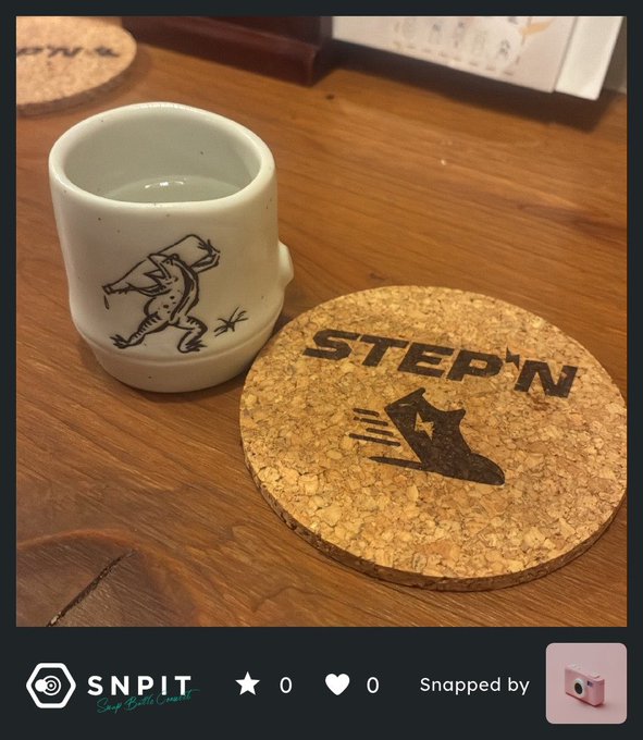 The image features a white cup with a black design, possibly of an animal or cartoon character, placed next to a cork coaster that has the word "stepn" written on it. The objects are positioned on top of a wooden table.