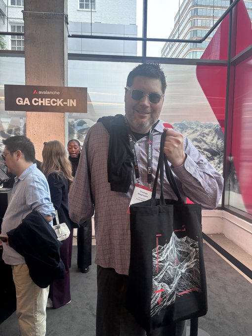 A smiling man with sunglasses and a lanyard holding a tote bag at an 'Avalanche GA CHECK-IN' booth during a conference.