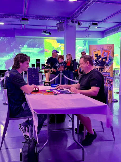Two people are seated across from each other at a table playing a board game in a room with purple lighting, with a sign for 'FAZENDO GAME' visible in the background.