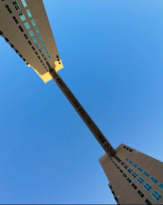 Low-angle architectural photograph looking up at two beige buildings connected by a skybridge against a clear blue sky.