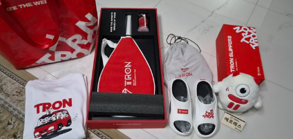 A collection of TRON-branded merchandise and packaging, including a red and white bag, slippers, a drawstring pouch, a face mask, and a small plush mascot character.