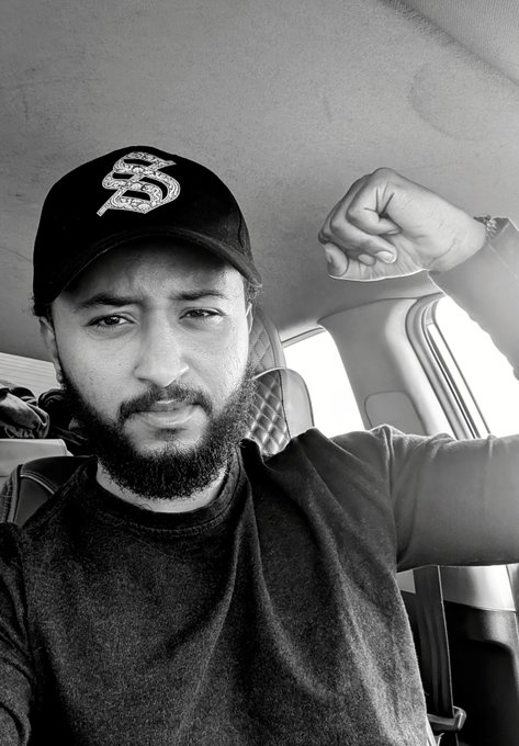 A black and white selfie of a bearded man wearing a 'Sonic' cap, sitting inside a vehicle with his arm raised.