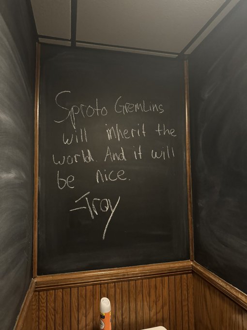 A handwritten message in white chalk on a black chalkboard that reads, "Sproto Gremlins will inherit the world. And it will be nice. -Troy," with an orange spray can resting below.