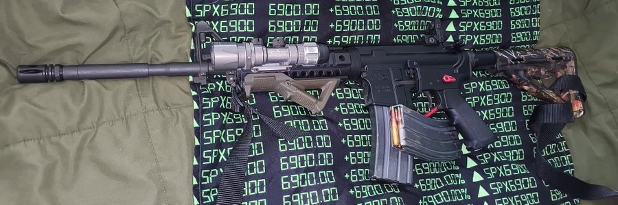 An AR-15 style rifle equipped with a suppressor, flashlight, foregrip, and camouflage sling, laid on a green bag with a stock market ticker-style fabric showing repeated 'SPX6900 +6900.00' and upward green triangles.