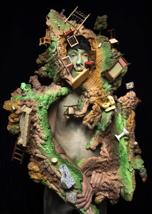A surreal artistic image showing a person's face and bare torso enveloped in an elaborate miniature landscape featuring rocky terrain, trees, ladders, and tiny furniture pieces against a black background.