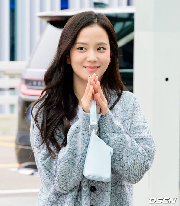 A young woman with long wavy dark hair wearing a gray cardigan and carrying a light blue handbag stands smiling with her hands clasped together in a greeting gesture, photographed by OSEN.