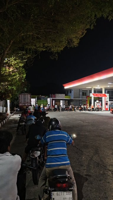A line of motorcyclists waiting at a gas station at night, with a license plate reading 'K 6277 ADF' visible on the nearest bike.