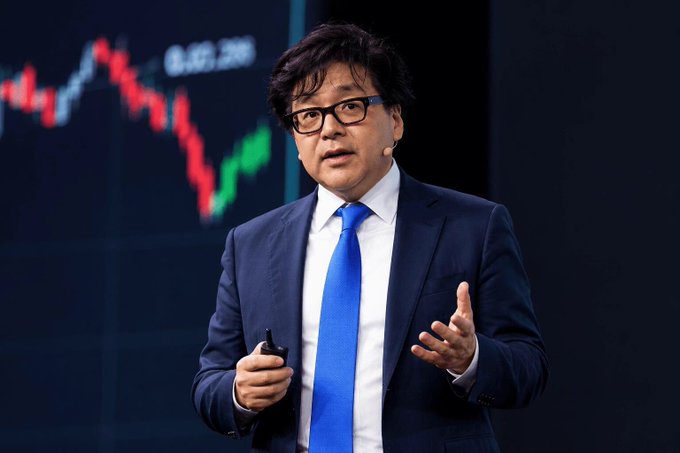 A middle-aged man wearing a navy blue suit, white shirt, and bright blue tie speaks while gesturing with his hand, standing in front of a dark screen displaying a red and green stock graph.
