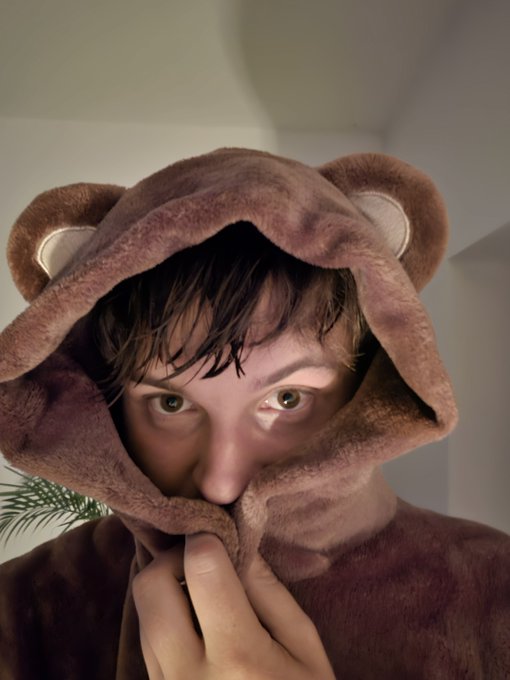 A young man wears a brown fuzzy bear-ear hooded garment, pulling it up over the lower half of his face with one hand.