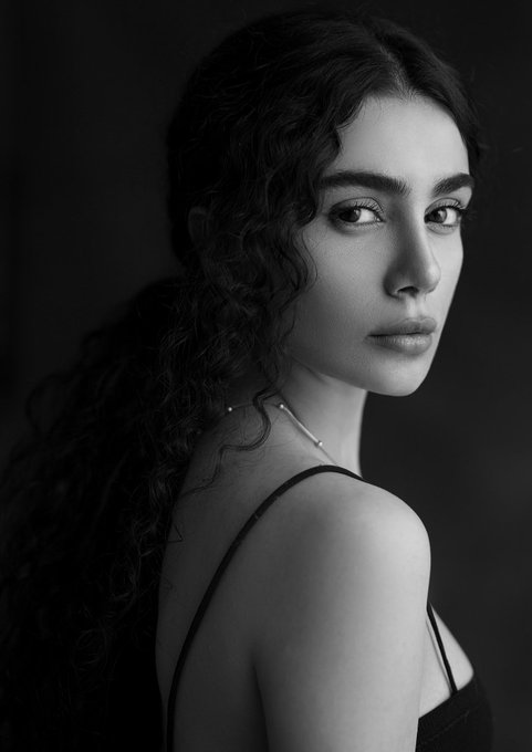 A black and white portrait of a young woman with curly hair looking over her shoulder, wearing a thin-strapped top and a delicate necklace.