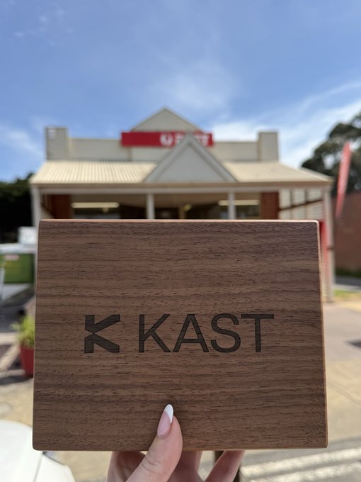 A hand holds a wooden box engraved with the 'K KAST' logo in front of a building with a red sign.