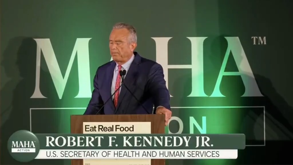 A man in a blue suit and red tie speaking at a podium with the text 'Eat Real Food', labeled 'ROBERT F. KENNEDY JR. U.S. SECRETARY OF HEALTH AND HUMAN SERVICES' at the bottom in front of a green 'MAHA' backdrop.