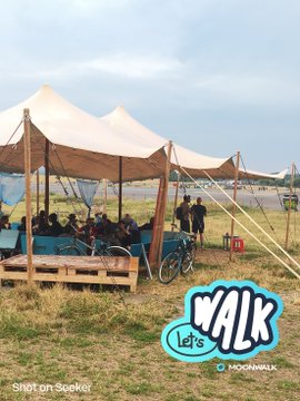 A photograph of people relaxing under a white tent at an outdoor event, with a 'Let's Walk' Moonwalk logo in the corner, taken on a Motorola phone.