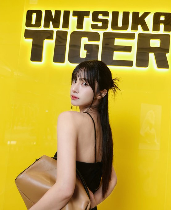 A young woman with long dark hair poses with her back turned, looking over her shoulder in front of a bright yellow wall featuring illuminated 'ONITSUKA TIGER' signage.