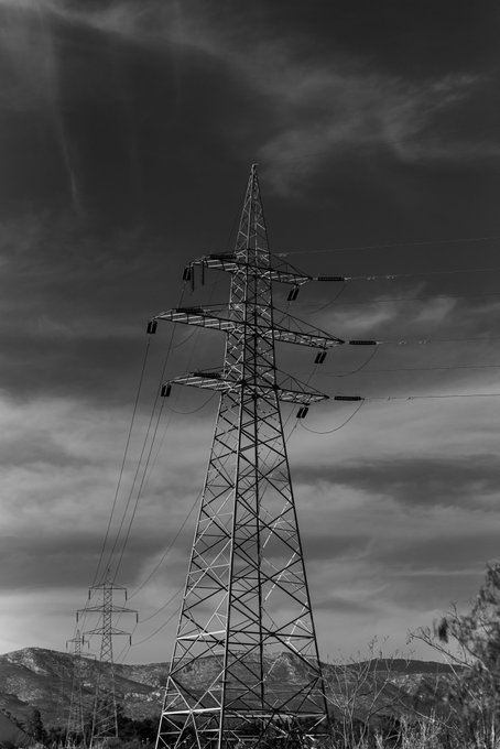 A black-and-white photograph of a tall metal electricity transmission tower against a dramatic cloudy sky, with a smaller tower and hills visible in the background.