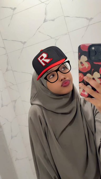 A selfie of a woman wearing a grey hijab, glasses, and a black baseball cap with a red 'R' logo, holding a phone with a cherry-printed case.