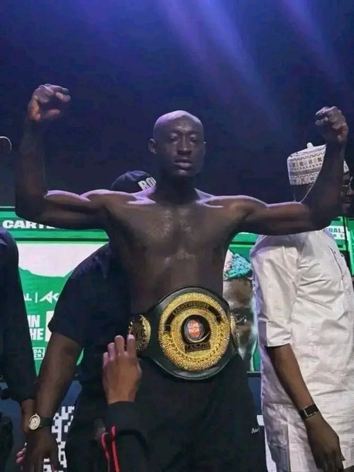 A shirtless, muscular bald man poses with both arms flexed while wearing a large championship belt, surrounded by people at what appears to be a sporting event.
