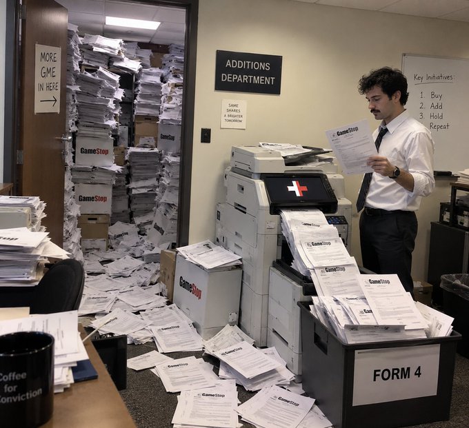 An office scene with a man surrounded by massive stacks of GameStop paperwork, signs reading 'MORE GME IN HERE,' 'ADDITIONS DEPARTMENT,' and a whiteboard listing 'Key Initiatives: 1. Buy, 2. Add, 3. Hold, 4. Repeat.'