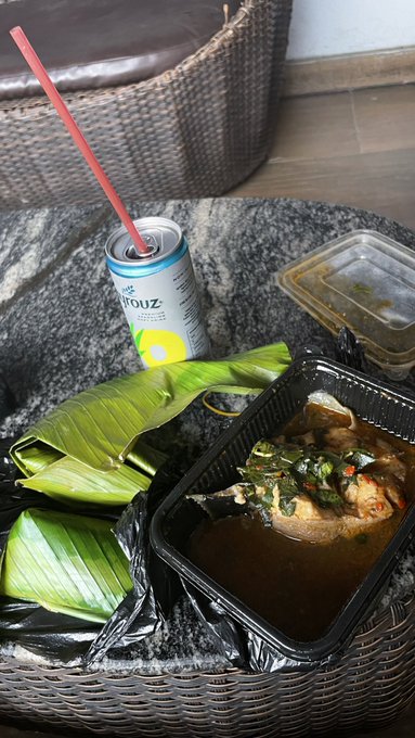 A takeout meal served on a banana leaf inside a black plastic container with curry/sauce, accompanied by a Stouz canned beverage with a red straw, sitting on a woven table.