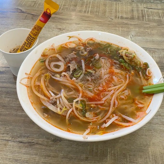 A bowl of spicy noodle soup with bean sprouts, meat, and vegetables sits next to a Trung Nguyen G7 coffee stick on a wooden table.