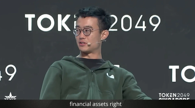 A man with glasses wearing a green Nike hoodie speaks on stage at TOKEN 2049 Singapore, with the subtitle 'financial assets right'.