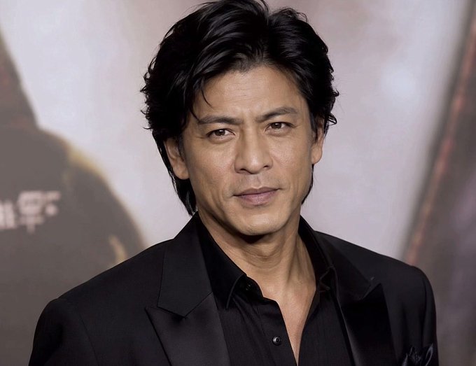 A man with dark, wavy hair wearing a black suit jacket over a black shirt poses at what appears to be a media event.