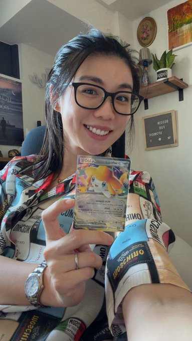 A smiling woman with glasses wearing a comic-print shirt holds up a Japanese Pokémon trading card featuring Jirachi.
