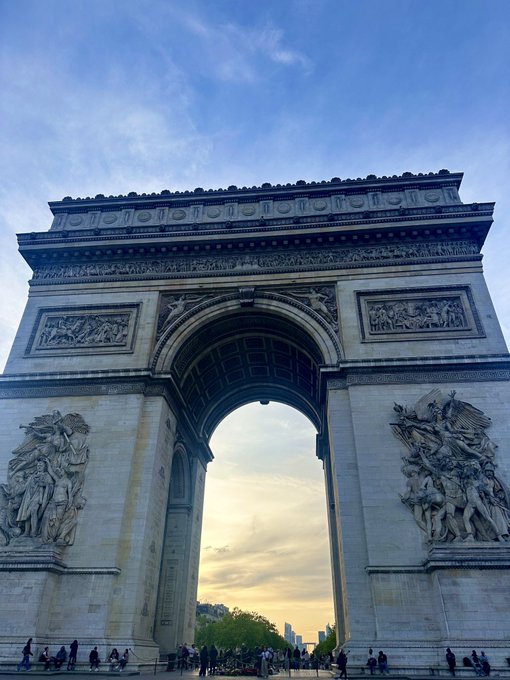 The Arc de Triomphe in Paris photographed at sunset, showing its ornate sculptural reliefs with tourists visible at its base.