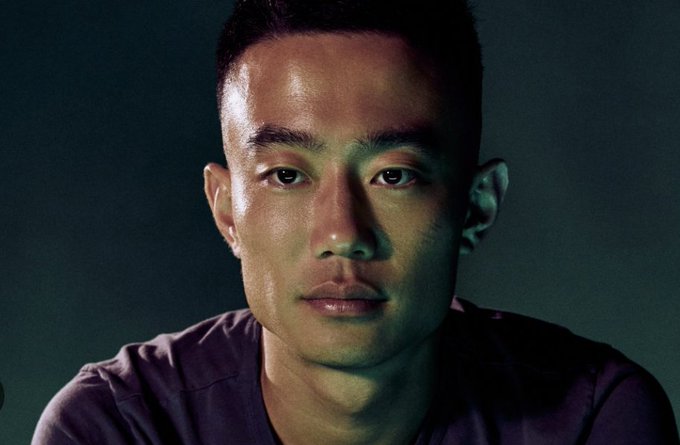 The image features a man with short hair wearing a purple shirt, looking directly at the camera. The background appears to be black, which contrasts with the man's light skin tone. There are no texts or other objects visible in the image.