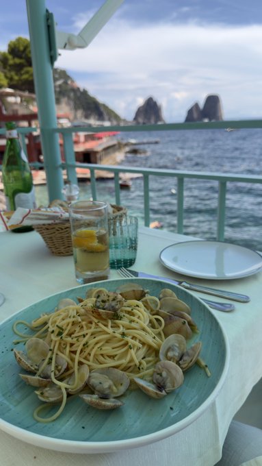 A plate of spaghetti alle vongole served at a seaside restaurant, with lemon water and a view of rocky sea stacks rising from the ocean in the background.
