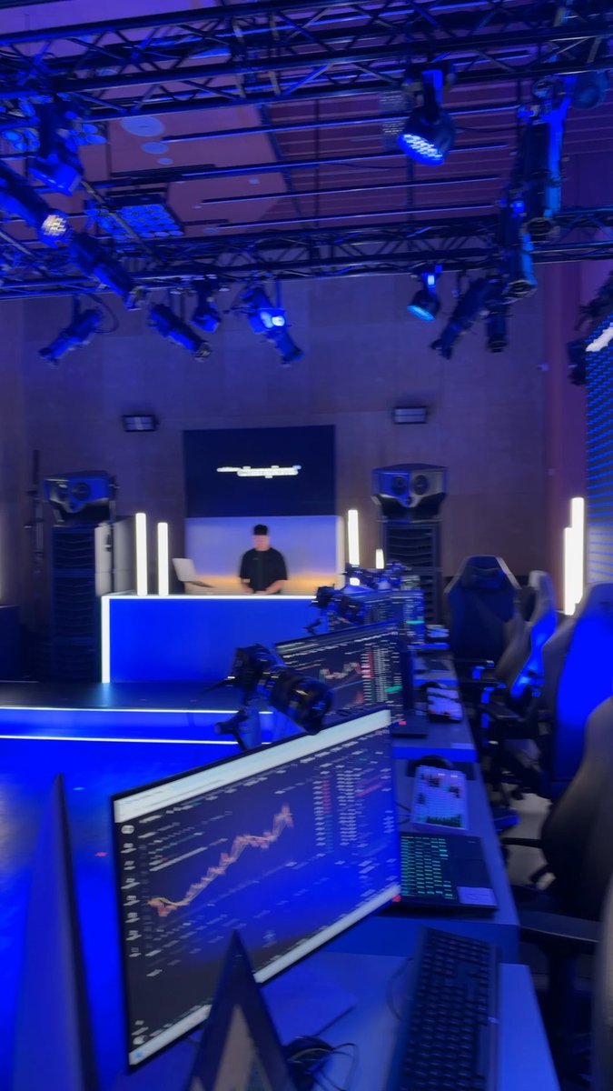A first look at Coinbase's trading competition battle station, featuring a blue-lit studio setup with multiple monitors displaying live trading charts and a host desk in the background. The professional staging with extensive stage lighting and gaming-style seats sets the scene for the upcoming competition.