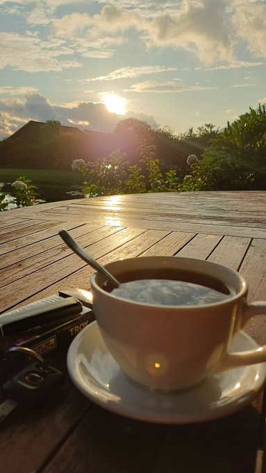 The image features a cup of coffee sitting on top of a wooden table with a spoon inside it. The sun is shining brightly through the window behind the cup, creating a warm glow throughout the scene. There are also some objects placed near the edge of the table, such as a car key and a cell phone.