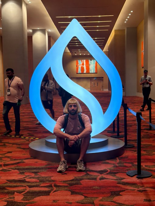 A person with a blonde bob wig sits cross-legged on the base of a large, illuminated blue Sui logo sculpture in what appears to be a conference venue.