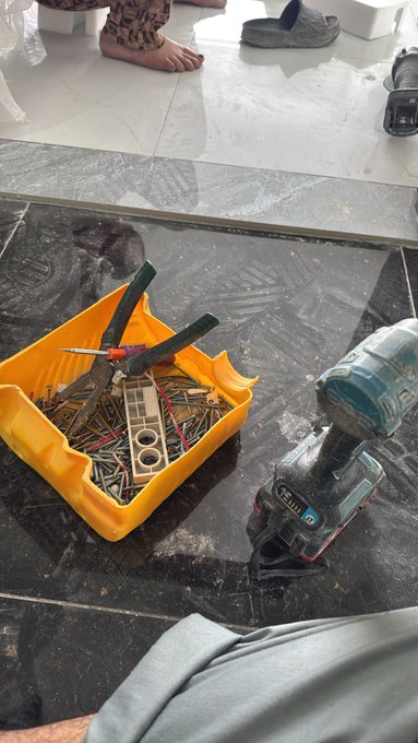 A construction site scene showing a yellow bucket filled with screws and tools, a power drill on a dark tile floor, with a person's bare feet visible in the background.