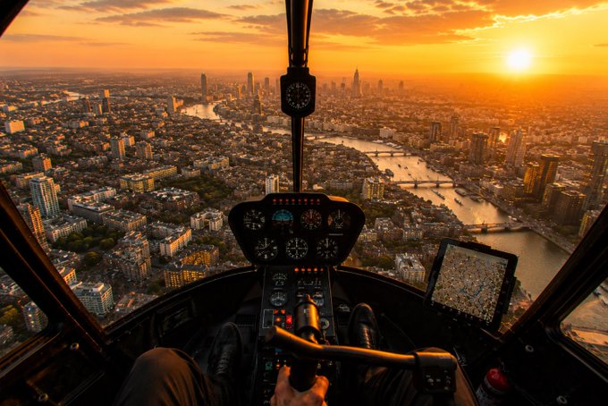 A first-person cockpit view from inside a helicopter flying over a sprawling city at golden hour, with a digital map display and analog instruments visible on the dashboard. The image mirrors the post's confident tone of steady progress and calm control, with the pilot's hand on the controls symbolizing the hands-off "no action required" approach while XRP and GM charts run their course.