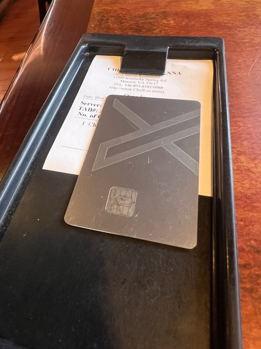 A black server check presenter folder sits on a wooden table, holding a document from 'CHELTENNA' and a grey card with a geometric 'Z' design and an EMV chip.
