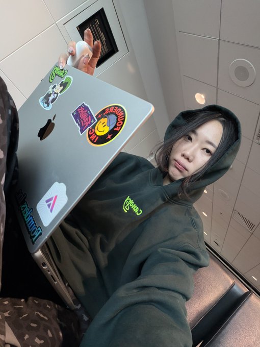 A young woman in an olive green hoodie takes a selfie with a MacBook covered in stickers, including a Smiley Face burger and logo stickers.