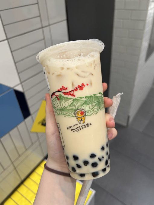 A hand holds a clear plastic cup of bubble tea with black tapioca pearls, decorated with a green wave design and text reading 'Give you a tea company'.