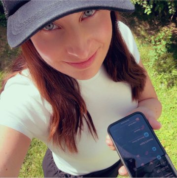 A selfie of a woman wearing a gray sun hat and white t-shirt, smiling while holding a smartphone outdoors, with what appears to be a trading app visible on the screen.
