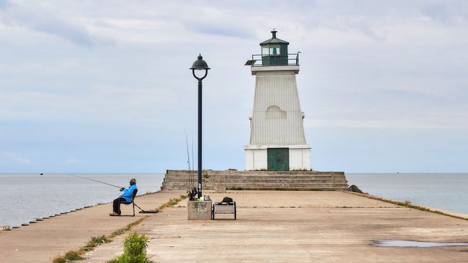 A person sits fishing on a pier in front of a small white lighthouse by the sea under a cloudy sky.