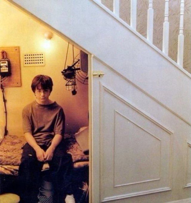 A young man with glasses sits somberly under a staircase on a bed, surrounded by a cluttered space with exposed wires and electronics, capturing a melancholic mood that mirrors the post about returning to live with parents after a failed investment trade.