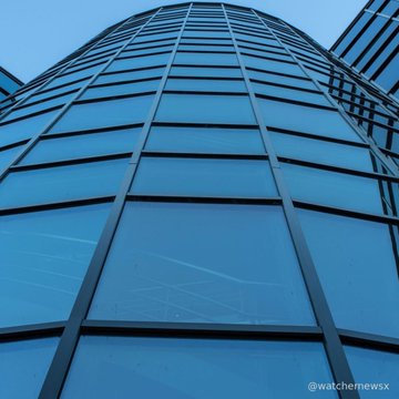 A low-angle photograph looking up the facade of a modern glass skyscraper with blue-tinted windows reflecting the sky, watermarked @watchnewsx.