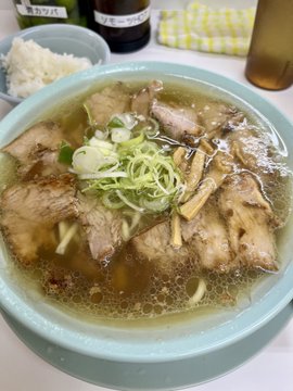 The image features a bowl of soup with meat, noodles, and vegetables such as onions and celery floating in water. The soup appears to be a beef noodle dish served in a blue bowl placed on a table. In the background, there are two bottles - one containing rice and another possibly filled with beer.