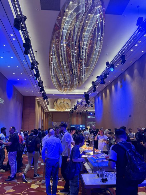An indoor shot of a crowded event or conference in a large ballroom with an ornate, spiraling golden chandelier hanging from the ceiling under blue lighting.